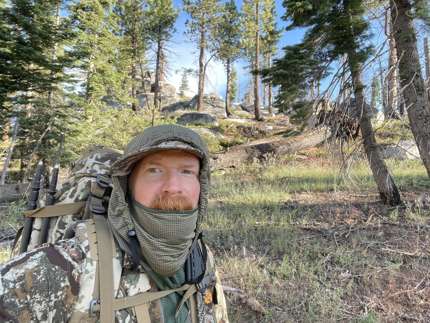 James Reed on a backcountry hunt, loaded pack and trekking poles over his shoulder, granite outcrop and pines behind him