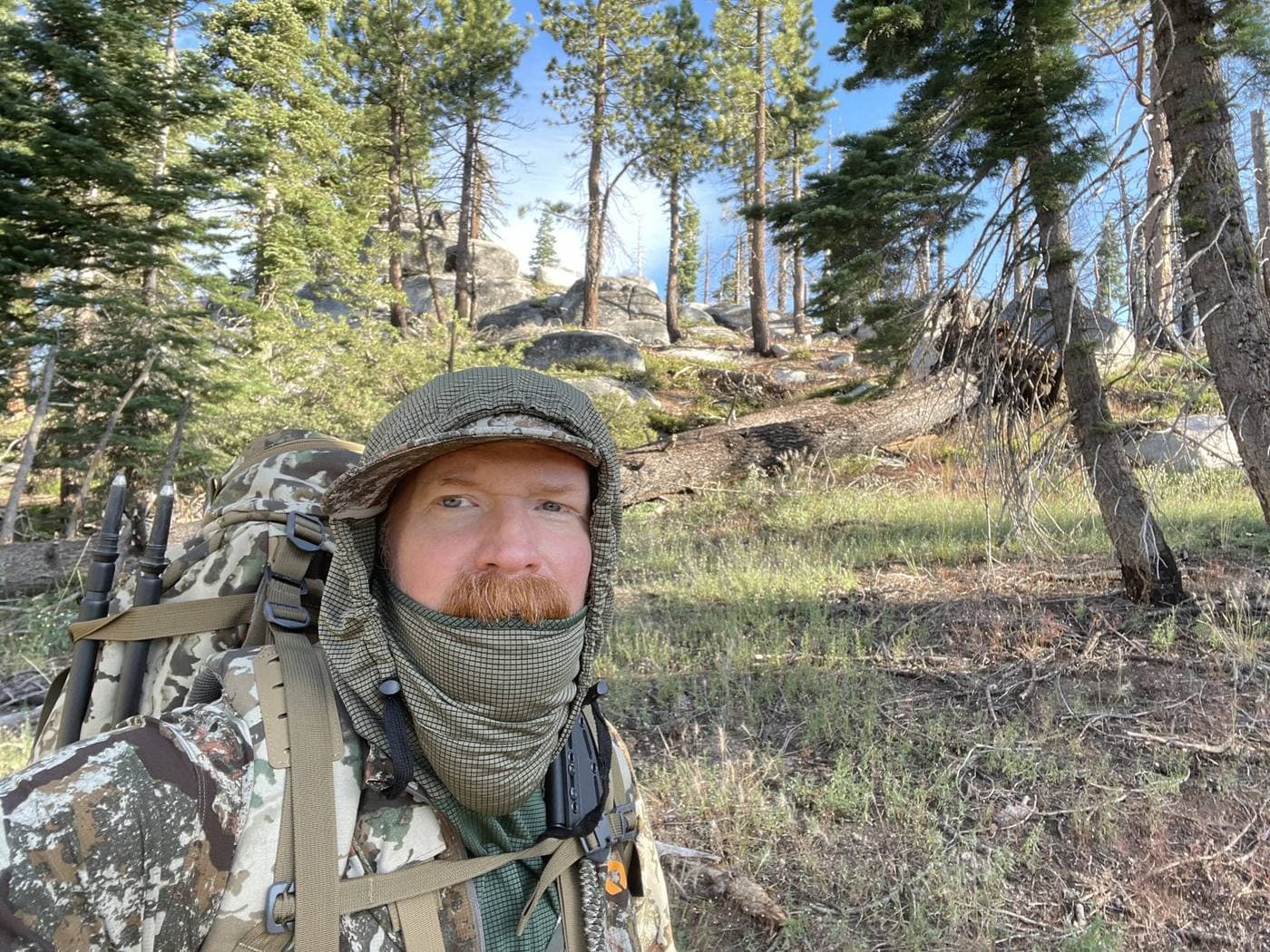 James Reed on a backcountry hunt, loaded pack and trekking poles over his shoulder, granite outcrop and pines behind him