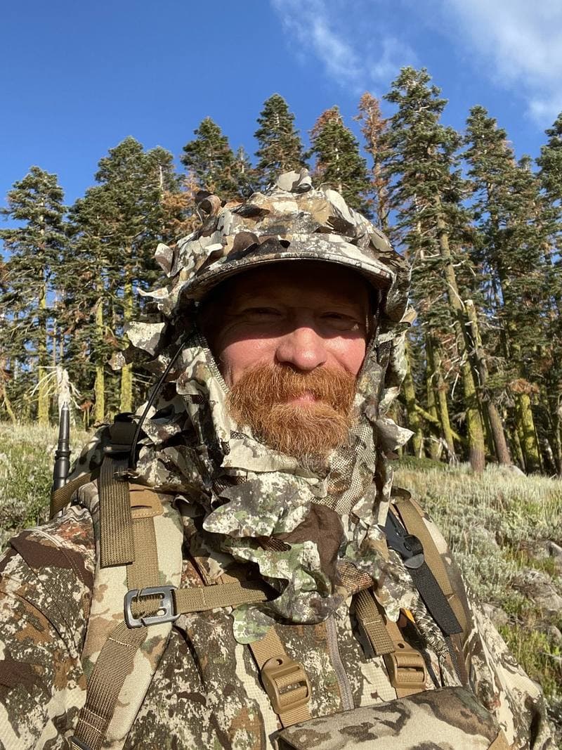 James Reed in a leafy camouflage hood and pack straps, red beard, tall conifers behind him