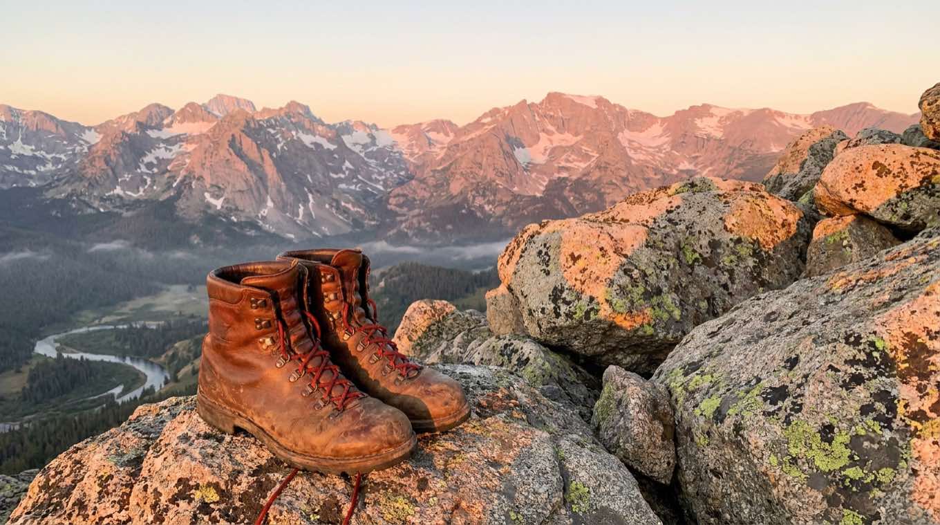 Worn leather mountain boots on lichen-covered granite above a river valley, alpenglow on the peaks behind